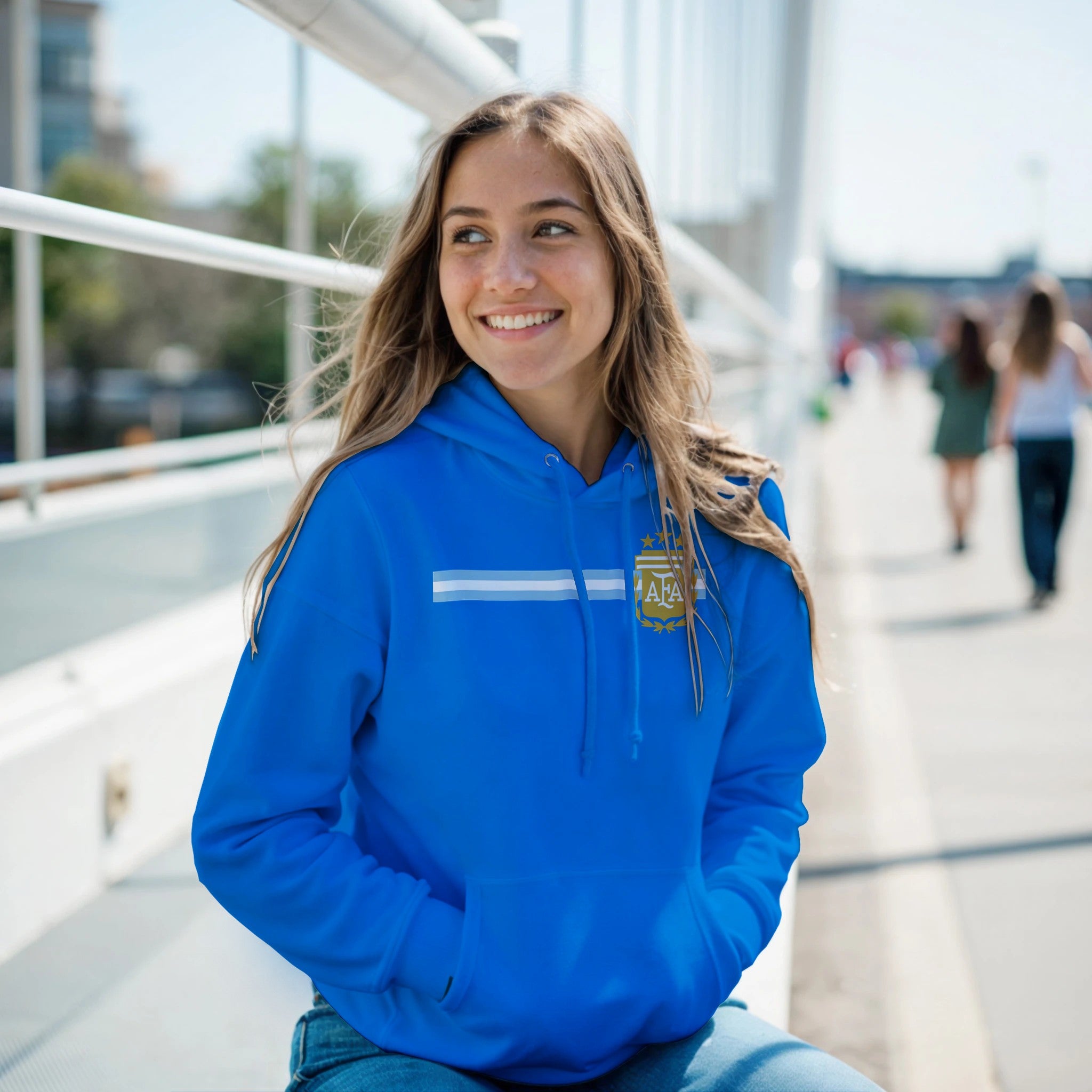 Woman wearing a blue hoodie with a logo, sitting outdoors on a sunny day.