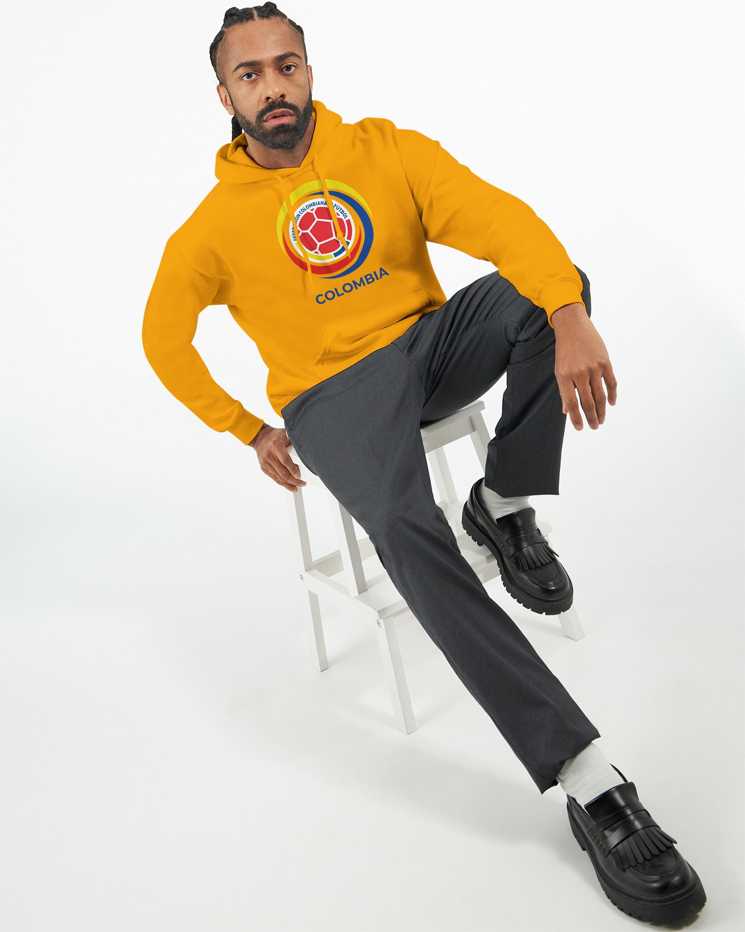 Person wearing a yellow Colombia hoodie, sitting on a white stool against a white background