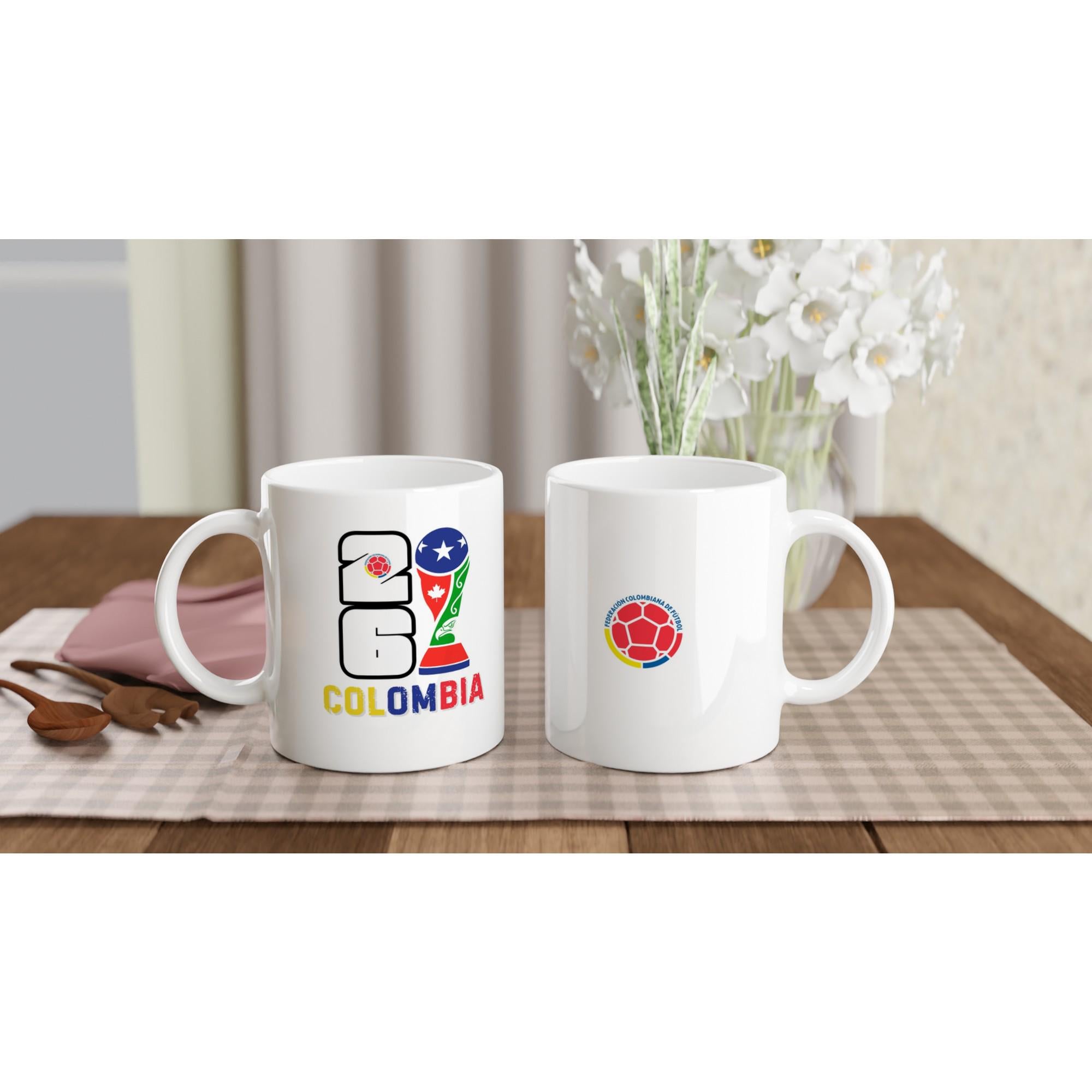 Two white mugs with Colombia-themed designs on a table with a checkered tablecloth and flowers in the background.